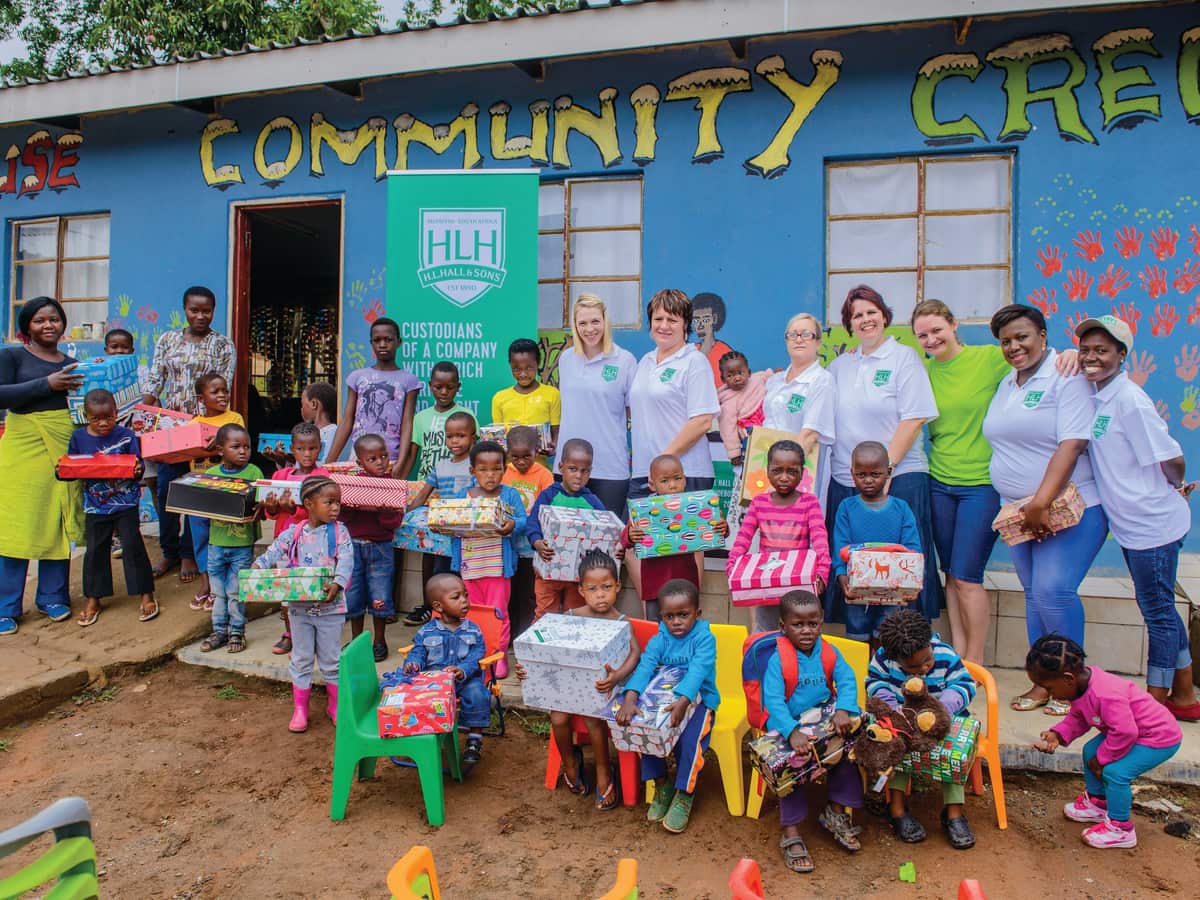 Halls staff in branded shirts and local children holding gift boxes outside a brightly painted community crèche